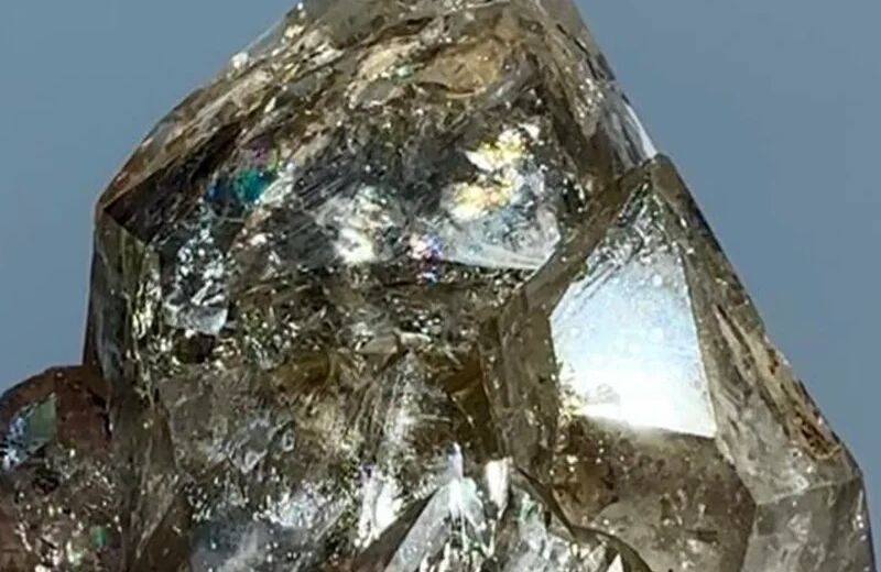 Close-up of a smoky Herkimer diamond with glassy faces and rainbow reflections against a blue-gray background